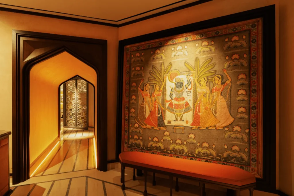 A hallway features a colorful Indian mural depicting figures and palm trees. Warm lighting, archways, and a red bench create an inviting atmosphere.
