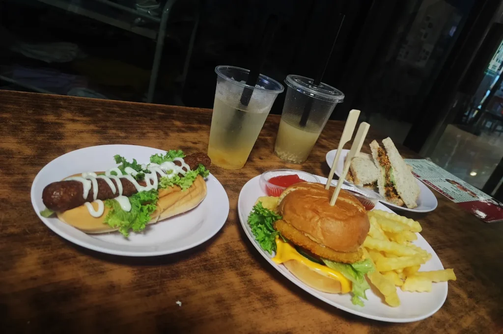 Plates with hot dog, cheeseburger, fries, and sandwich on a wooden table. Two lemon drinks with black straws sit beside the plates, creating a casual dining vibe.