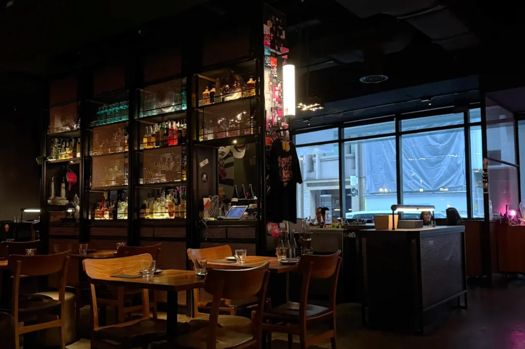 Cozy dimly lit bar with wooden tables and chairs. Shelves display various bottles. A large window reveals an urban street view, creating an intimate ambiance.