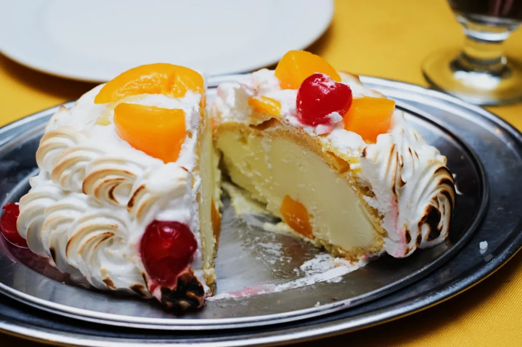A halved Baked Alaska dessert on a metal tray. The outer layer is toasted meringue, adorned with peach slices and cherries, revealing creamy interior.