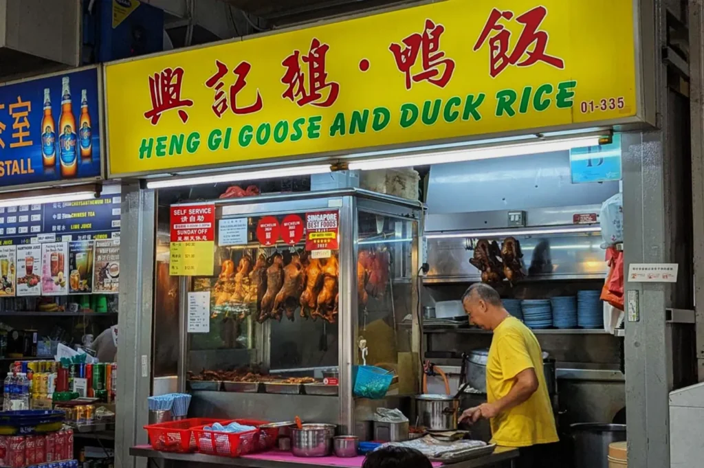 Market food stall sign "Heng Gi Goose and Duck Rice." Displayed roast ducks hang inside. A vendor in a yellow shirt prepares food, suggesting a bustling atmosphere.