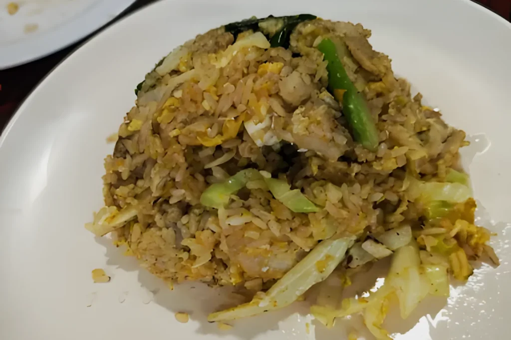 Fried rice served on a white plate, featuring bits of egg, sliced green vegetables, and pieces of cabbage. The dish has a savory, homemade feel.
