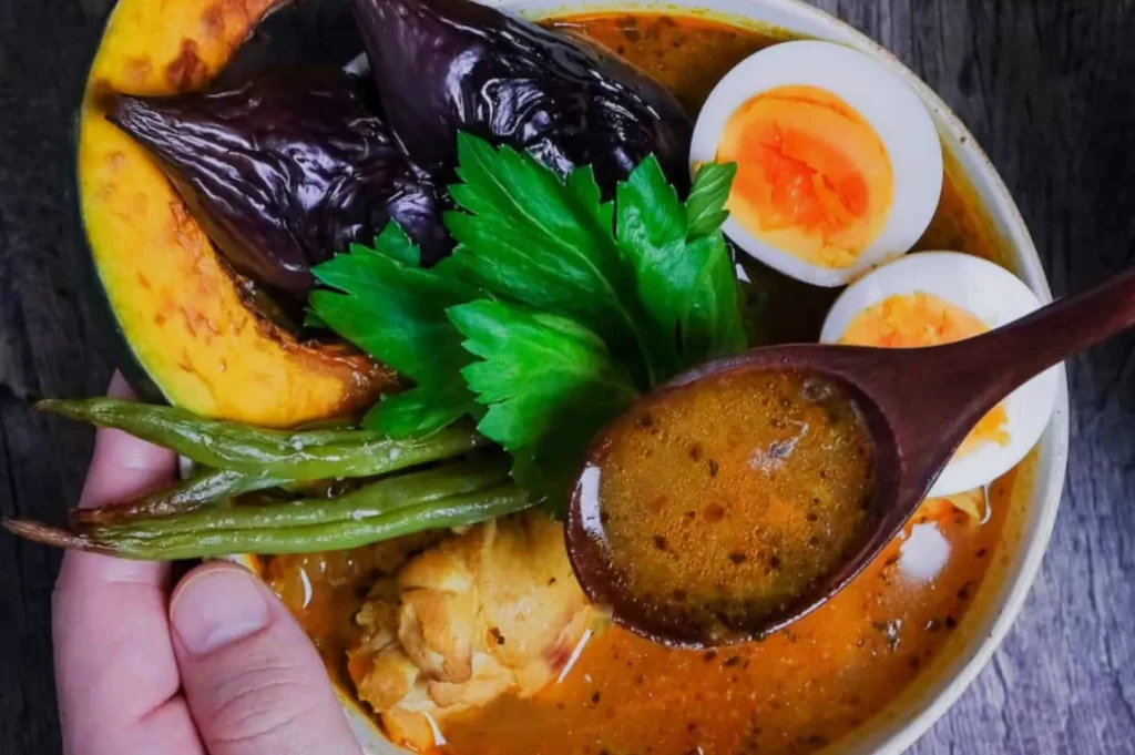 A vibrant bowl of curry features a rich broth in a wooden spoon, boiled eggs, eggplant, green beans, squash, and cilantro, evoking warmth and comfort.