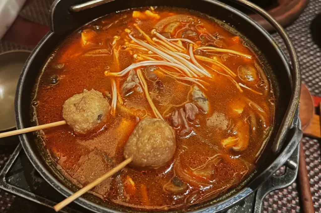 A steaming pot of rich, spicy soup with vibrant red broth. It features enoki mushrooms, meatballs on skewers, and assorted vegetables, exuding warmth.