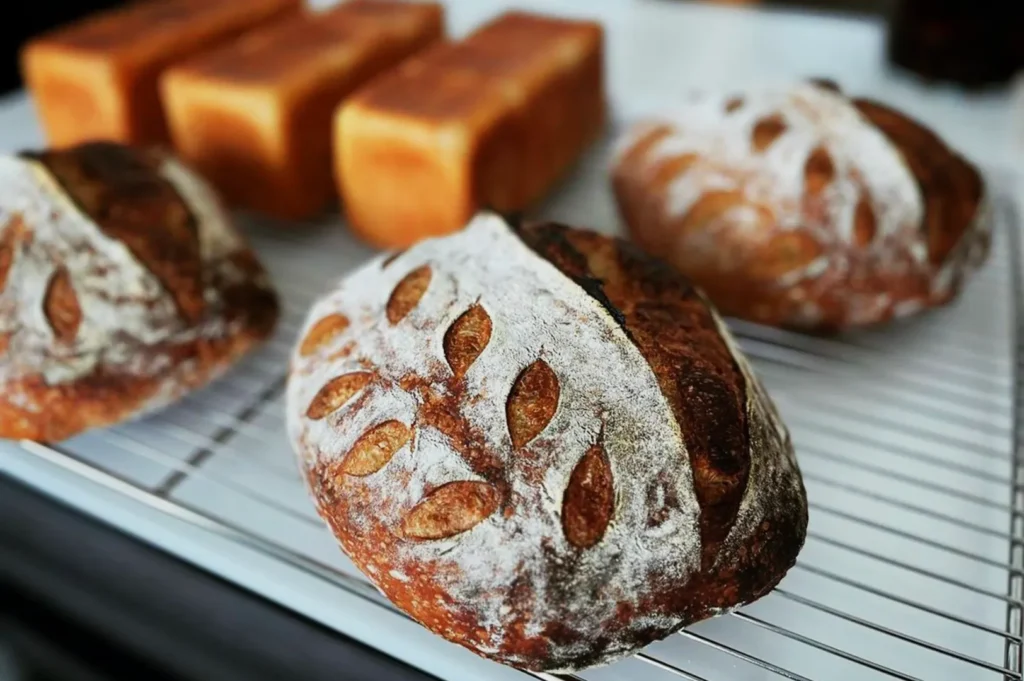Freshly baked round loaves with leaf patterns and rectangular bread on a cooling rack. The setting suggests a warm and inviting bakery.