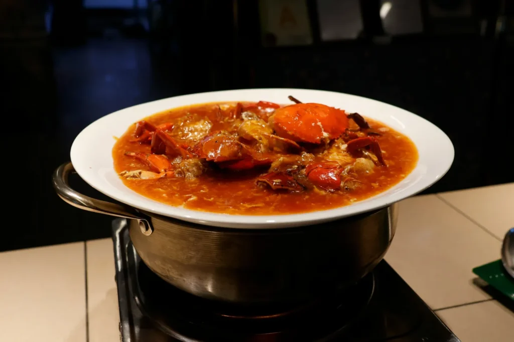 A steaming bowl of chili crab in rich, reddish-brown sauce sits on a hot pot. The scene is warmly lit, evoking a cozy dining ambience.