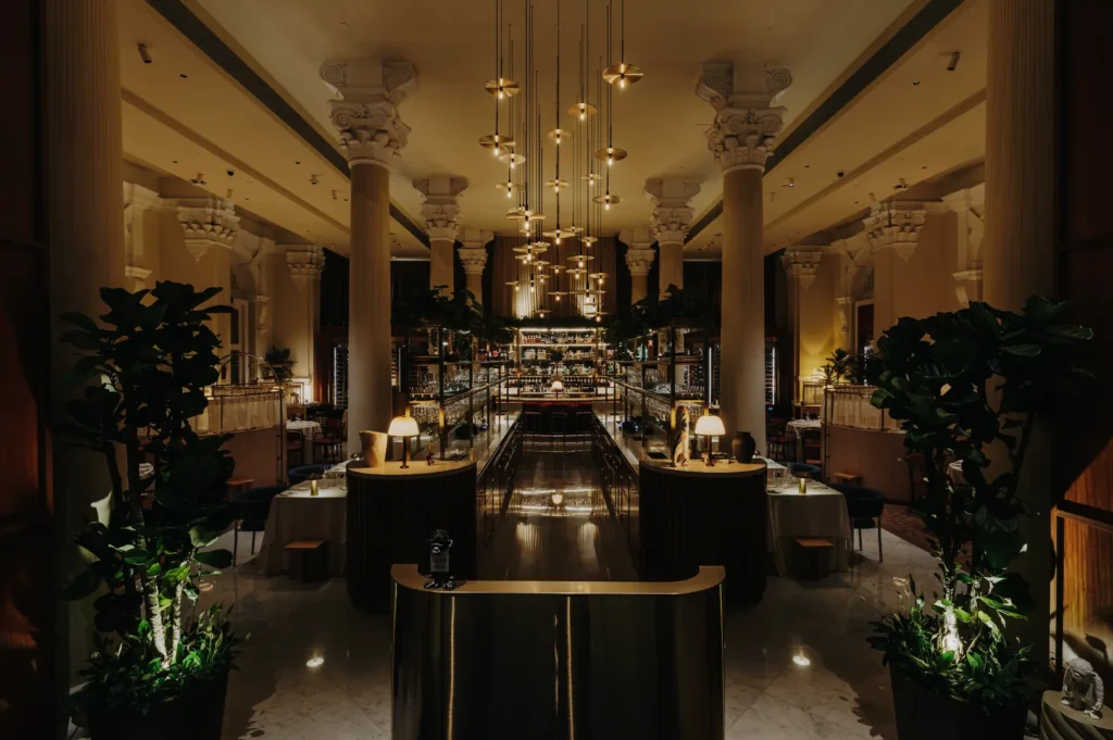 Elegant restaurant interior with dim lighting, tall columns, and modern chandeliers. Plush seating and lush plants create a luxurious atmosphere.