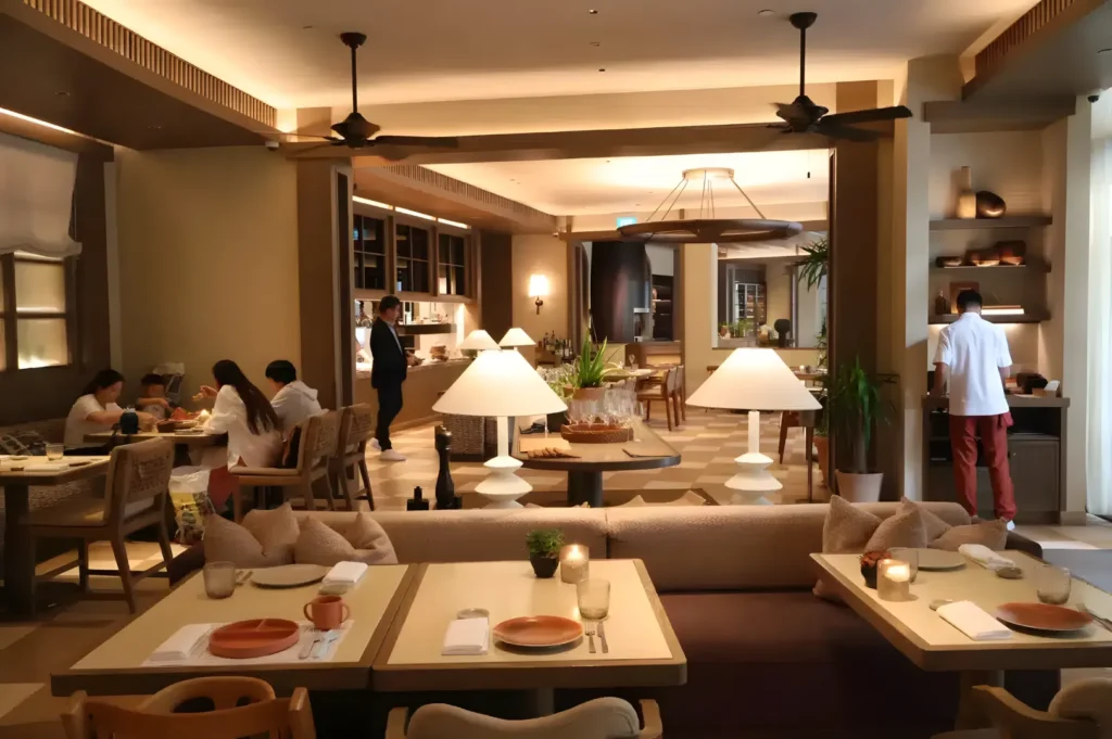 Modern restaurant with warm lighting, wooden tables, and cozy seating. Patrons are dining, staff is serving, creating a relaxed, inviting ambiance.