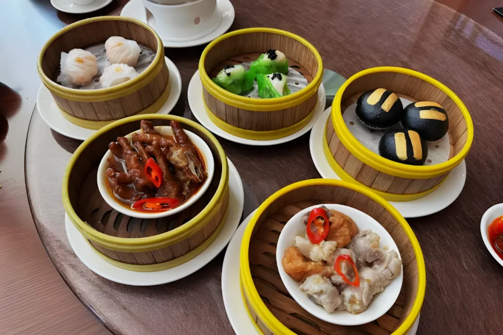 A table features bamboo steamers with various dim sum dishes: shrimp dumplings, green dumplings, black buns, chicken feet with chili, and dumplings with red chili slices, creating a vibrant and inviting scene.