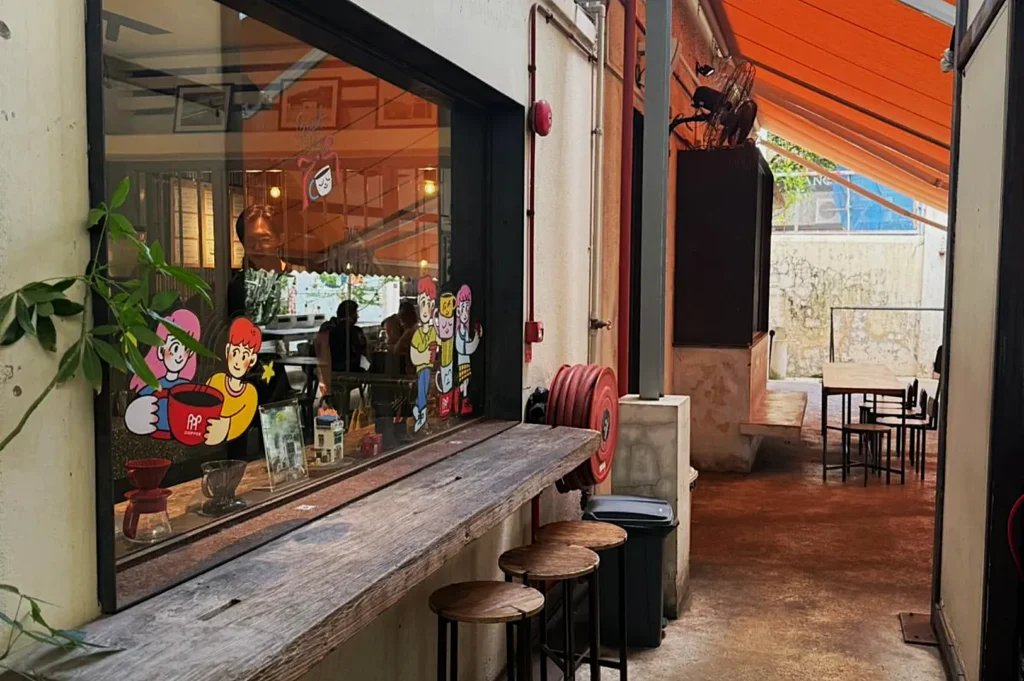 Narrow cafe alley with orange canopy, wooden counter, and stools. Cartoon figures on a window add a playful touch. Cozy and inviting atmosphere.