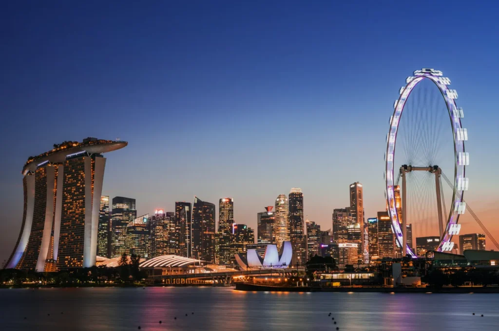 Singapore skyline at dusk, featuring Marina Bay Sands on the left, a lit Ferris wheel on the right, and bright city skyscrapers reflecting on the water.