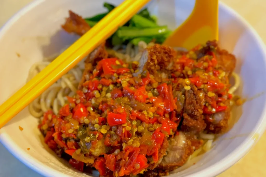 A bowl of noodles topped with crispy fried chicken covered in spicy red chili sauce, accompanied by green vegetables. Yellow chopsticks and a spoon rest on the bowl, conveying a vibrant and appetizing tone.