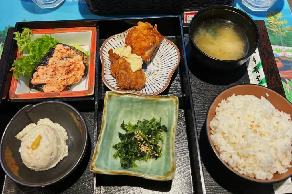A Japanese meal set on a black tray with rice, miso soup, grilled fish with pink sauce, fried chicken, potato salad, and seasoned greens.