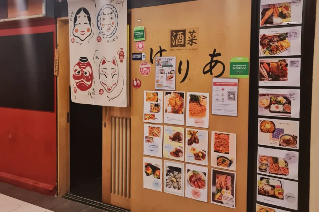 Japanese restaurant entrance with traditional curtain featuring painted faces. Menu items with vibrant food photos displayed on the wooden wall, creating a welcoming atmosphere.