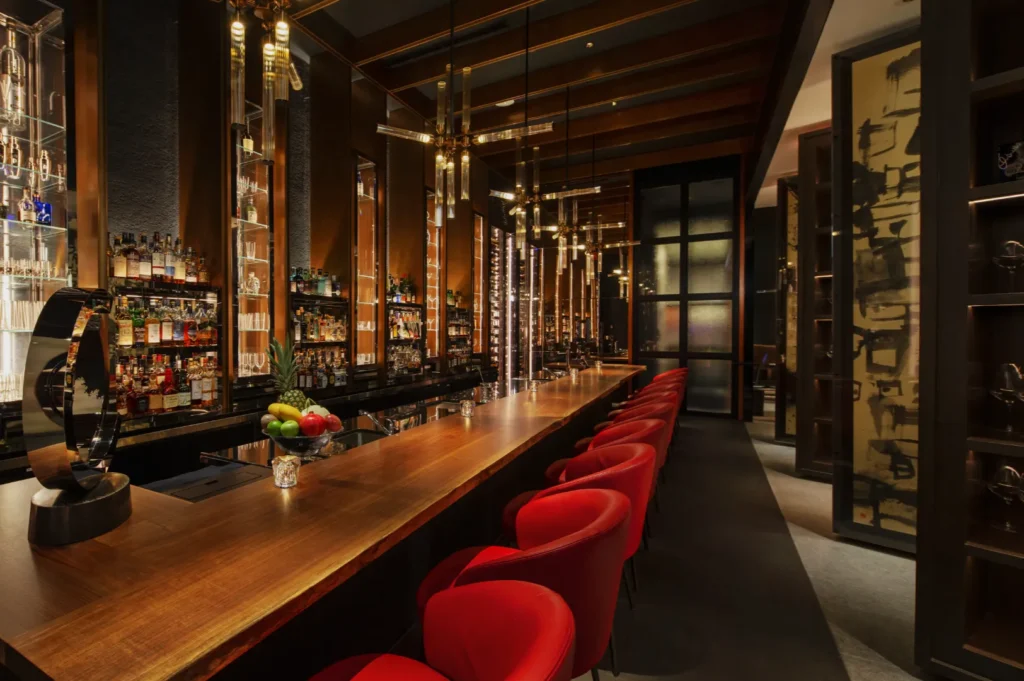 Elegant bar interior with a long wooden counter, red stools, and hanging lights. Shelves display various bottles. A vase of fruit adds color. Warm ambiance.