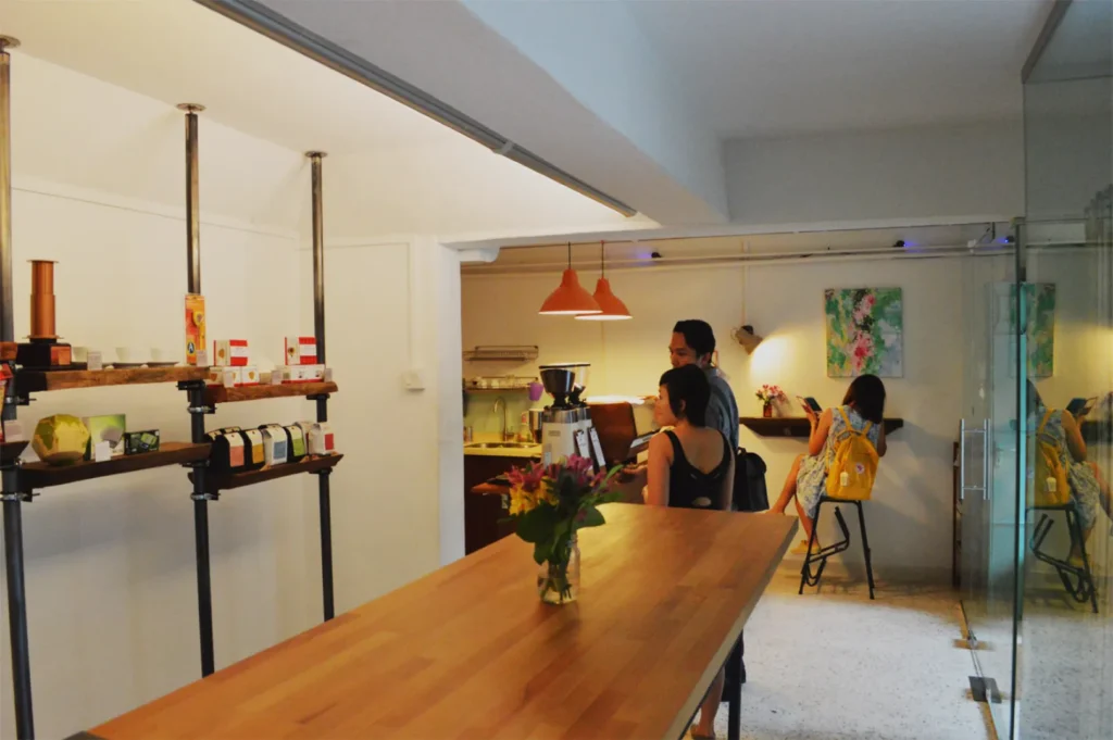 Cozy cafe with wooden counters, pendant lights, and shelves displaying coffee products. Two people order while another sits reading, creating a relaxed vibe.