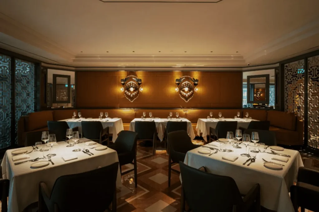 Luxurious restaurant interior with elegantly set tables, white tablecloths, and plush chairs. Warm lighting and patterned walls create a sophisticated ambiance.