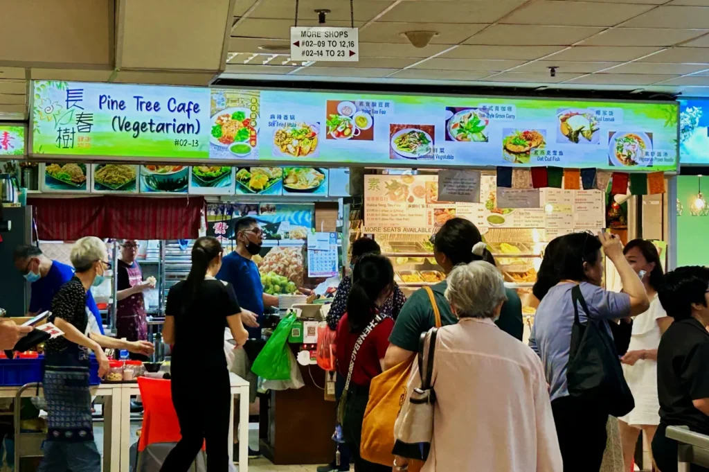 People line up at Pine Tree Cafe, a bright vegetarian food stall, with colorful menu boards overhead, conveying a lively and busy atmosphere.