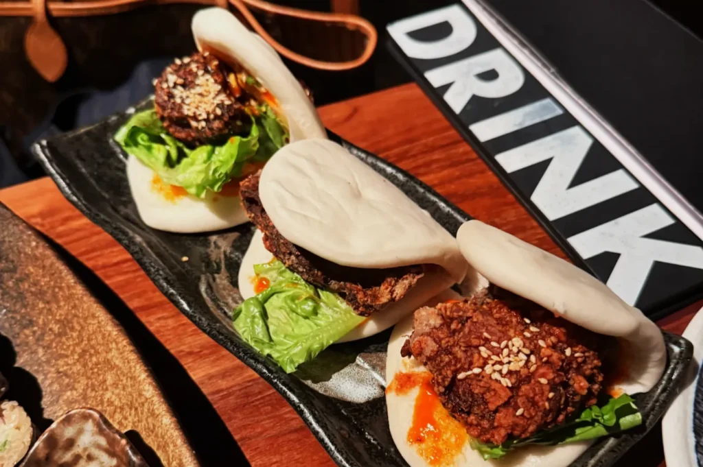 Three steamed bao buns are filled with crispy fried toppings, lettuce, and drizzled with sauce. They rest on a black plate beside a "DRINK" sign.