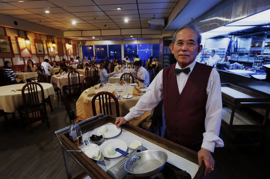 An older waiter in a burgundy vest and bow tie stands by a cart with cooking tools in a warmly lit, busy restaurant filled with dining patrons.
