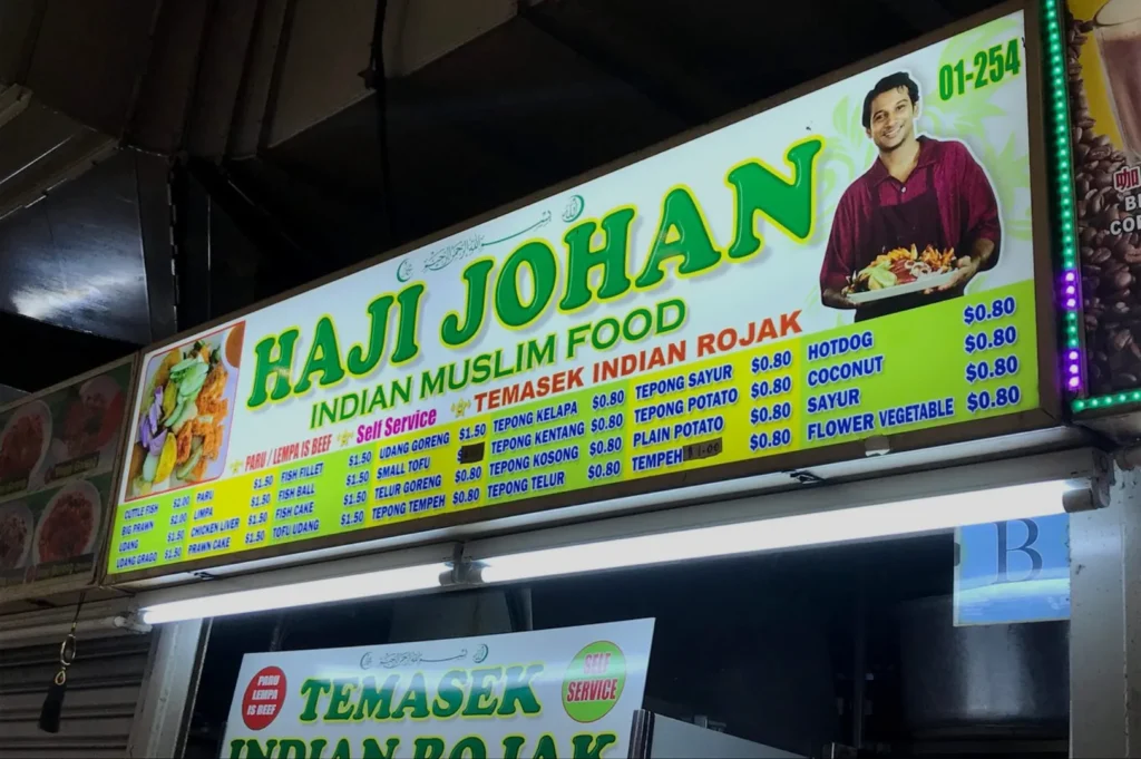 Bright sign displaying "Haji Johan Indian Muslim Food" with a smiling man holding a dish. Menu items and prices in bold; colorful and inviting tone.