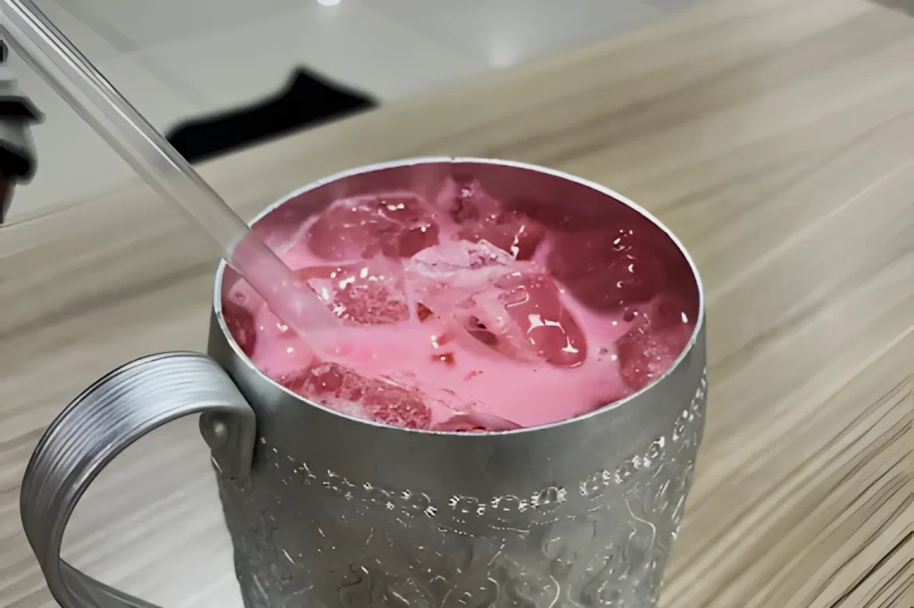 A silver cup filled with ice and pink liquid sits on a light wooden table. A straw is inserted into the drink, evoking a refreshing, inviting feel.