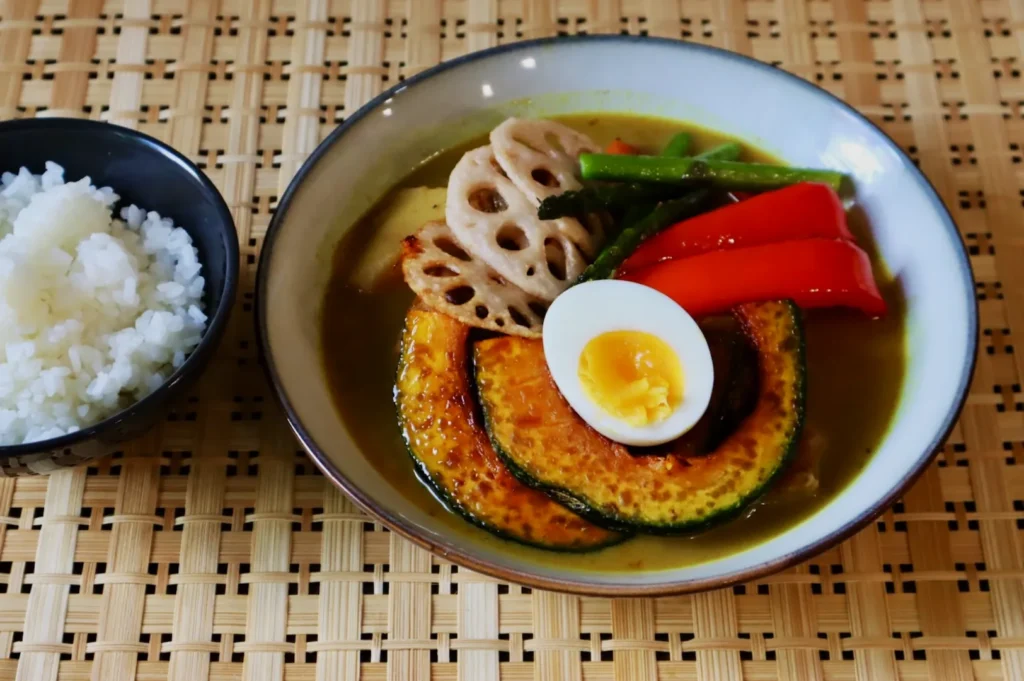Bowl of Japanese curry with sliced kabocha, red pepper, lotus root, green beans, half a boiled egg, and a side of white rice on woven mat.