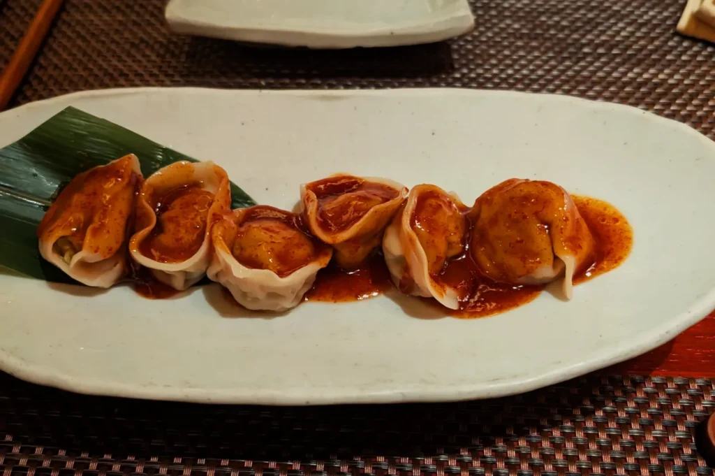 Six dumplings drizzled with a rich, dark sauce are presented on an oval white plate, garnished with a green leaf, creating an inviting and savory visual.