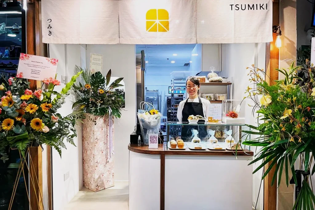A cheerful bakery shop adorned with vibrant flower arrangements. A smiling person in an apron stands behind the glass counter displaying pastries.
