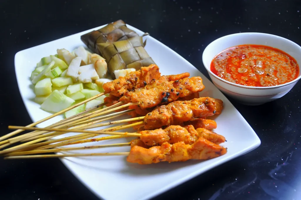 Grilled chicken satay skewers on a plate with cucumber and ketupat, paired with a bowl of spicy peanut sauce, set on a dark surface.
