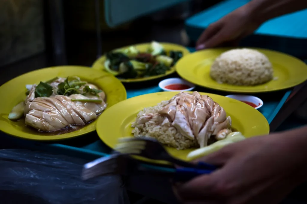 Yellow plates with Hainanese chicken rice, vegetables, and dipping sauce. A hand is serving the dishes, creating a casual, appetizing scene.