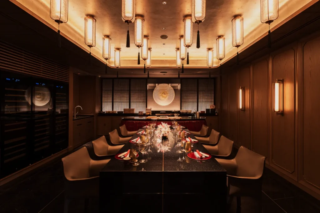 Elegant dining room with modern lighting, a long black table set with red-trimmed white plates, and cream chairs. Warm, inviting ambiance.