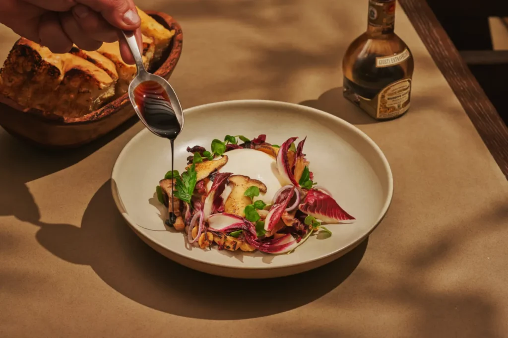 A hand drizzles balsamic vinegar from a spoon onto a gourmet salad of radicchio, mushrooms, and cheese. Warm bread and a vinegar bottle are nearby.
