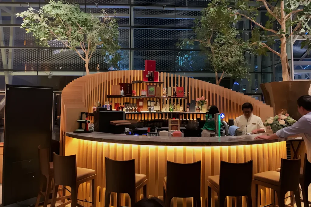 Contemporary curved bar with wooden accents and ambient lighting. Two bartenders and a patron interact. Tall indoor plants enhance the modern, inviting atmosphere.