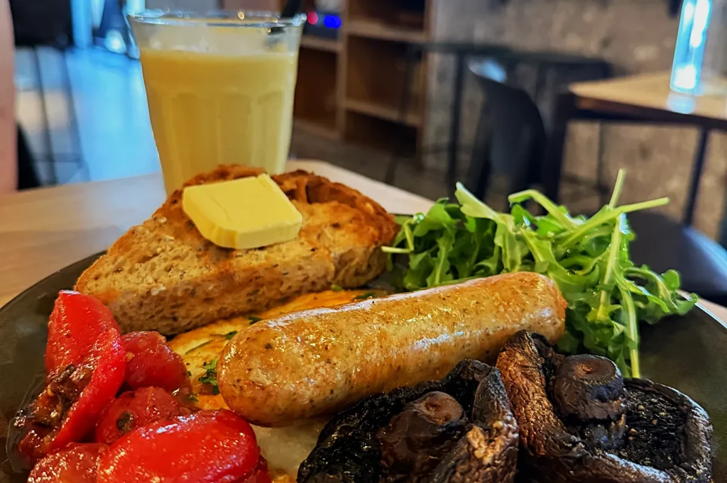 A hearty breakfast plate with toast topped with butter, grilled sausage, tomatoes, mushrooms, and arugula, accompanied by a glass of juice.
