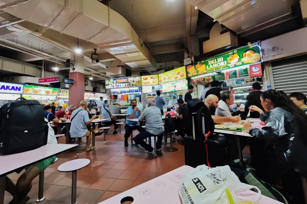 A bustling food court with diverse patrons seated at tables, enjoying meals. Bright menu signs are visible overhead, creating a lively, vibrant atmosphere.