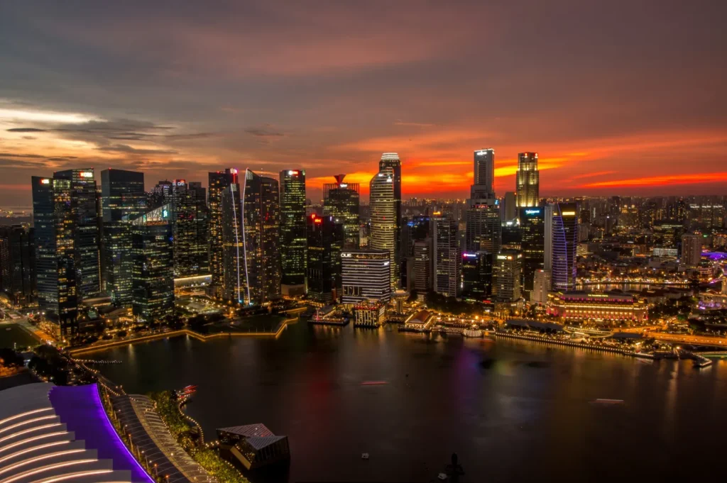 A vibrant city skyline at dusk with illuminated skyscrapers reflecting on calm water, against a dramatic orange-red sunset, conveying a lively, serene mood.