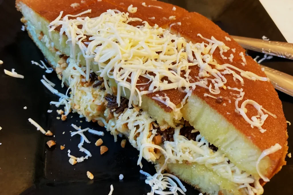 Close-up of a slice of Martabak, an Indonesian sweet pancake, topped with grated cheese. The filling includes chocolate and crushed nuts. Served on a black plate, creating a rich, indulgent appearance.