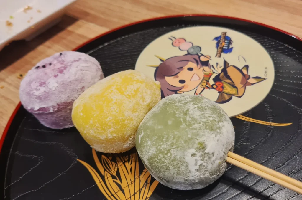 Three colorful mochi—purple, yellow, and green—sit on a black plate with a cute anime character design. The setting is light and playful.