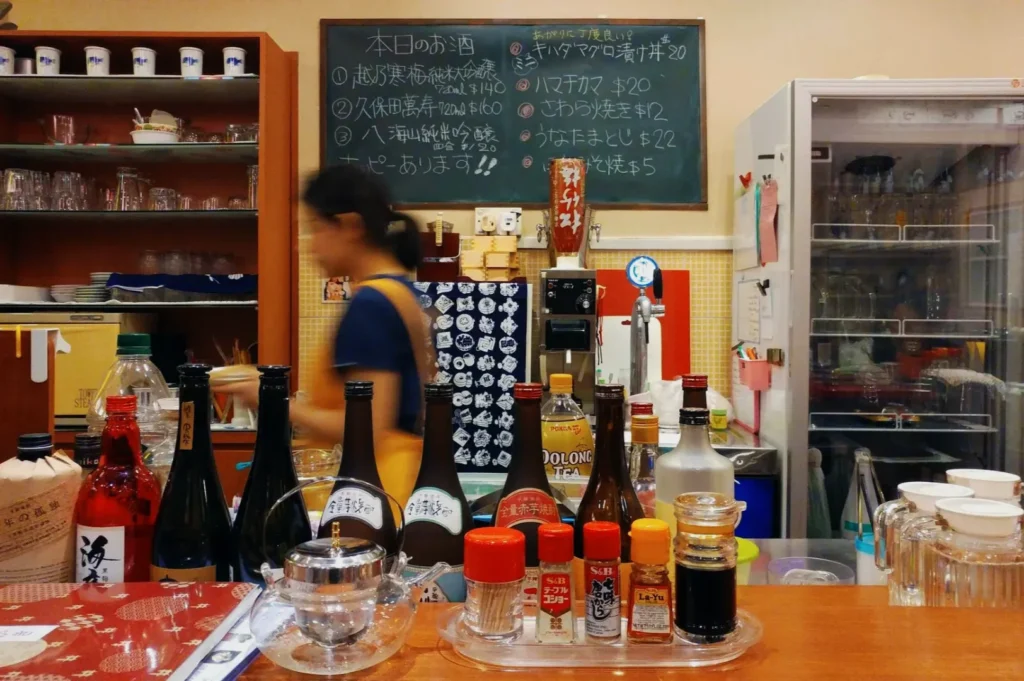 A woman in motion behind a bar counter with various sake bottles and condiments. A menu written in Japanese is on a chalkboard in the background.