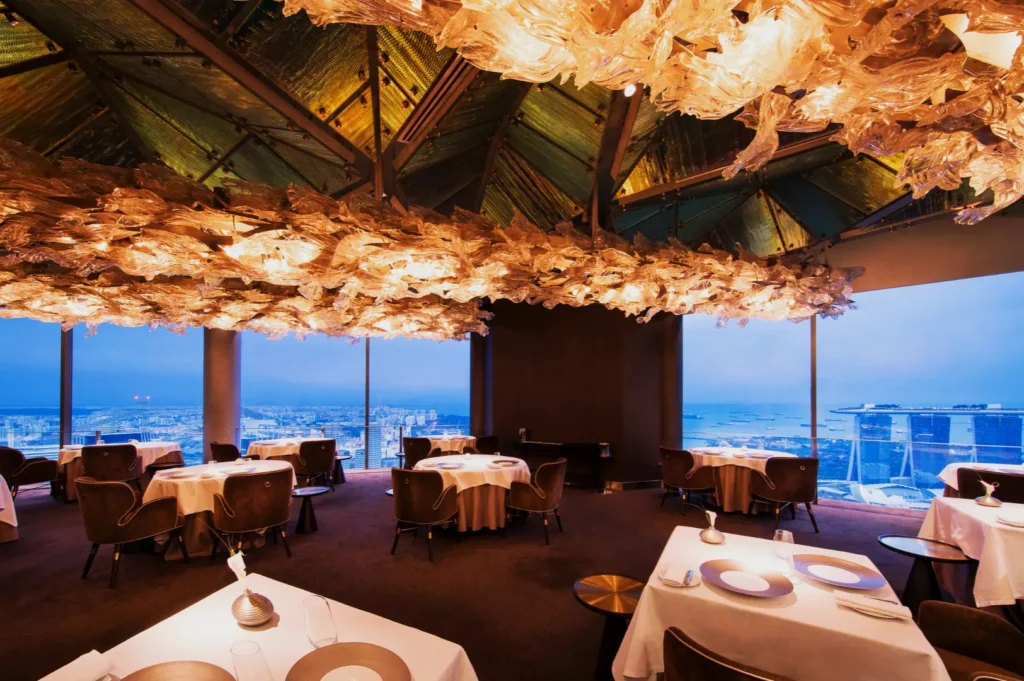 Luxurious restaurant with sculptural ceiling, ambient lighting, and panoramic city view. Elegant tables set with white linens create a serene atmosphere.