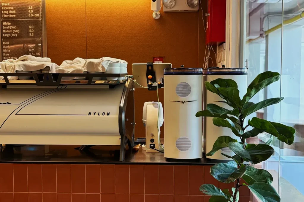 Modern cafe counter with a sleek white espresso machine labeled "NYLON," brown tiled surface, menu board, and potted plant. Warm, inviting ambiance.