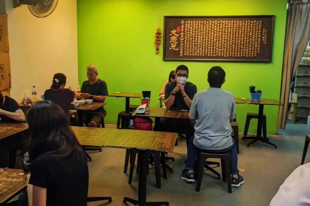 A cozy eatery with a lime green wall showcasing a large framed script. Patrons are seated at wooden tables, some conversing, one wearing a mask. Warm and relaxed atmosphere.