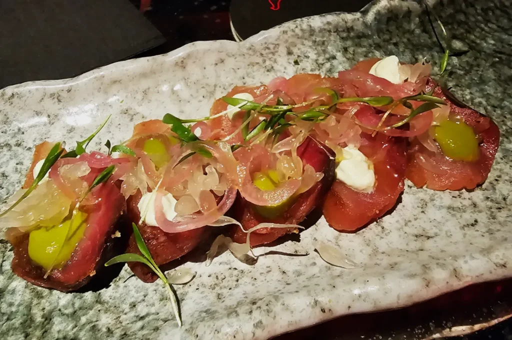 A plate of seared tuna topped with pickled onions, green sauce, and microgreens, artfully arranged on a textured gray ceramic dish.