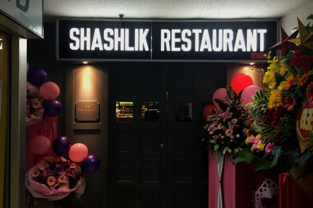 Entrance to Shashlik Restaurant, showcasing a dark door with a bright sign above. Decorated with colorful flower arrangements and balloons.