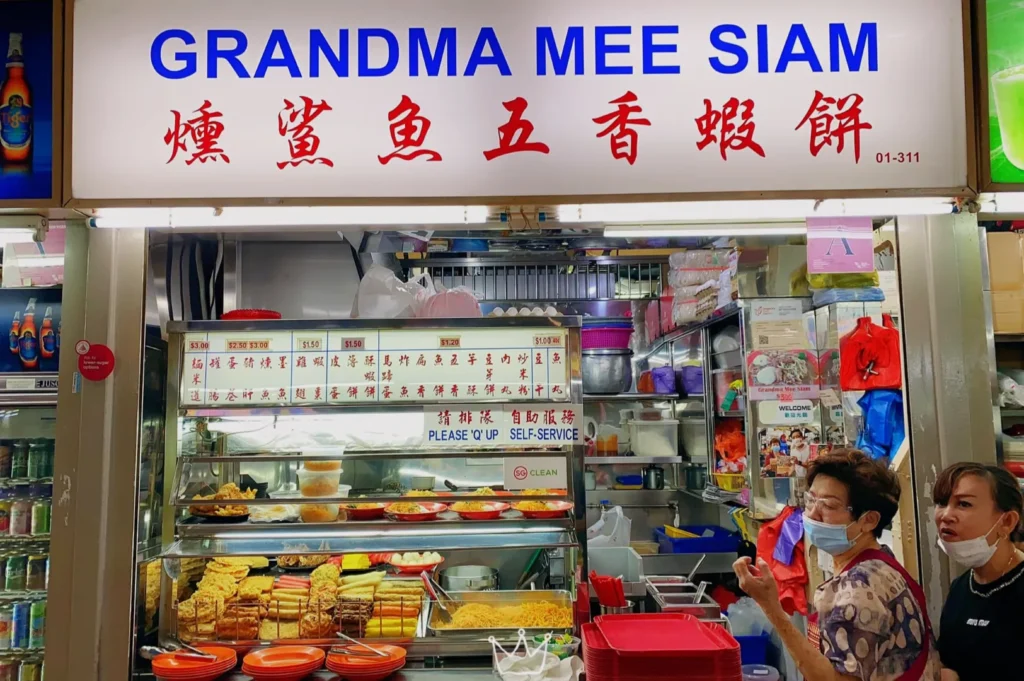 A hawker stall named "Grandma Mee Siam" displays a variety of colorful dishes. Two women, one wearing a mask, stand in front, creating a bustling, lively atmosphere.