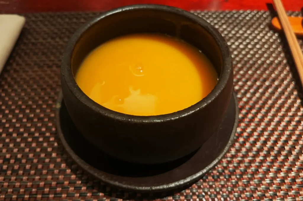 Dark bowl filled with bright orange soup on a textured mat, with chopsticks on the side. The scene conveys a warm, inviting culinary atmosphere.