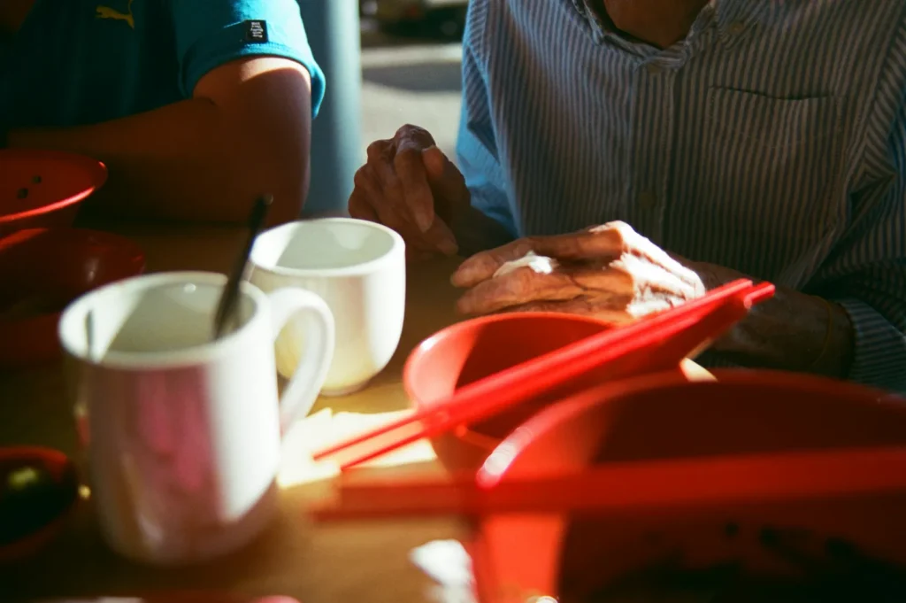 Two people sit at a table with red bowls and bright red chopsticks. Sunlight highlights an elderly hand touching the table, conveying warmth and serenity.