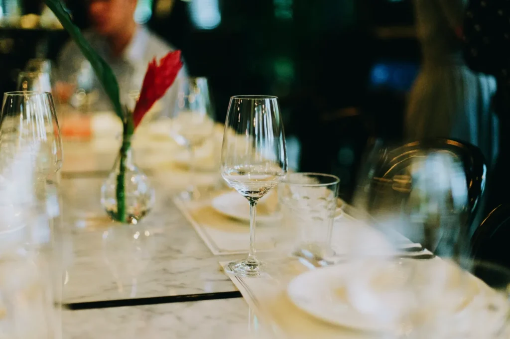 Elegant table setting with wine glasses, plates, and a vibrant red flower in a vase on a marble surface, creating a sophisticated dining ambiance.