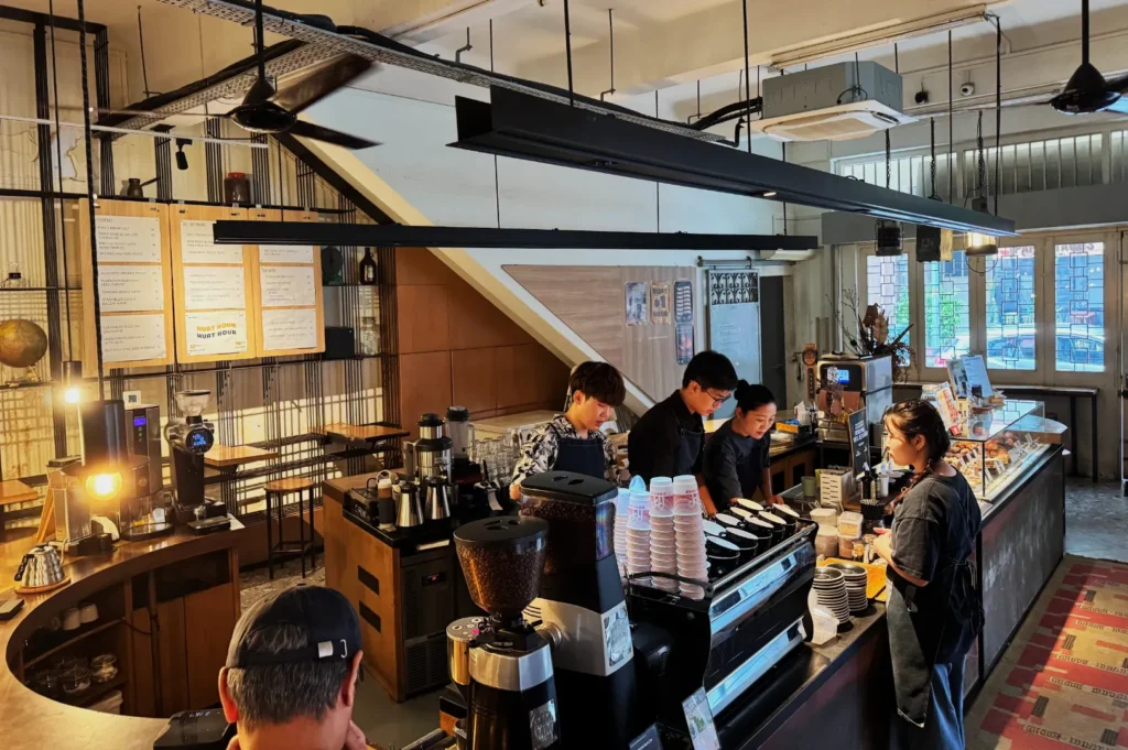 A cozy coffee shop with warm lighting and an industrial design. Three baristas prepare drinks behind the counter, with a customer waiting.
